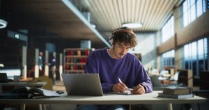Handsome Student Writing an Essay on Laptop Computer, Thinking About the Topic, Brainstorming, Finding Solutions. Young Stylish Man Studying in a Public Library, Listening to Lecture in Headphones