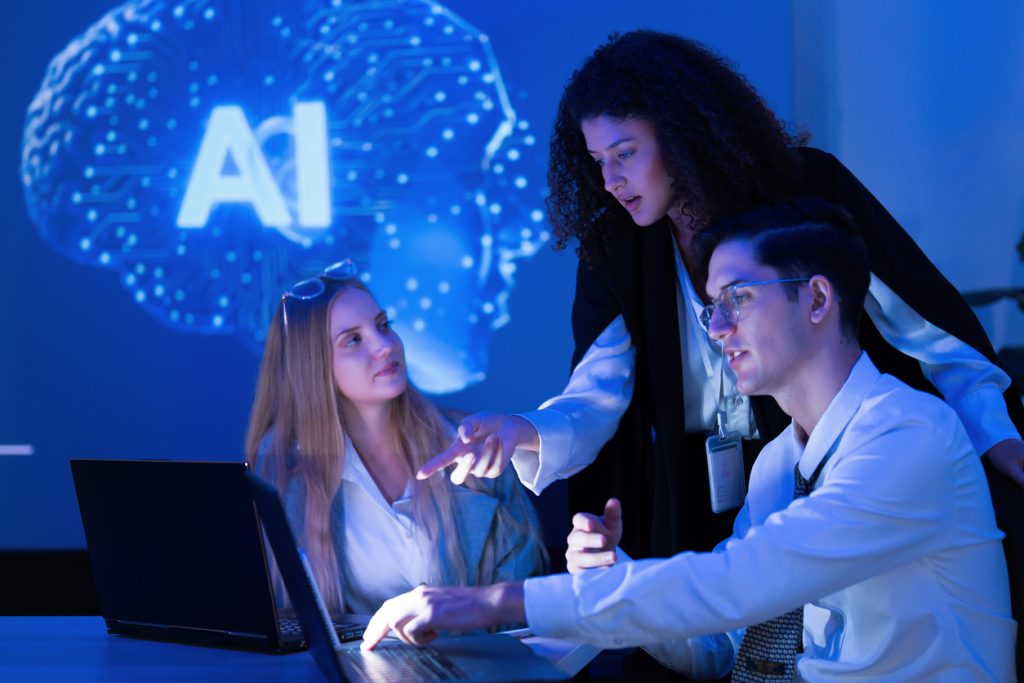 A group of three professionals engages in a discussion about an artificial intelligence project using a laptop. They work in a contemporary setting with blue ambient lighting.