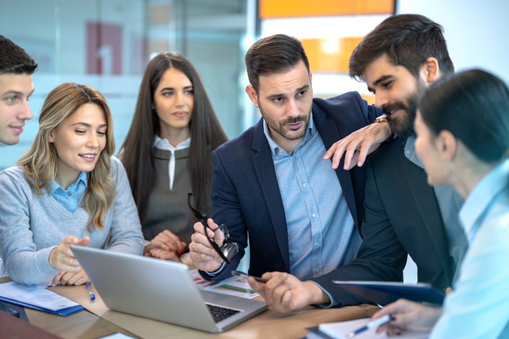 Handsome male CEO executive leader leaning on his colleague shoulders, explaining business strategy to his team colleagues at meeting in office.