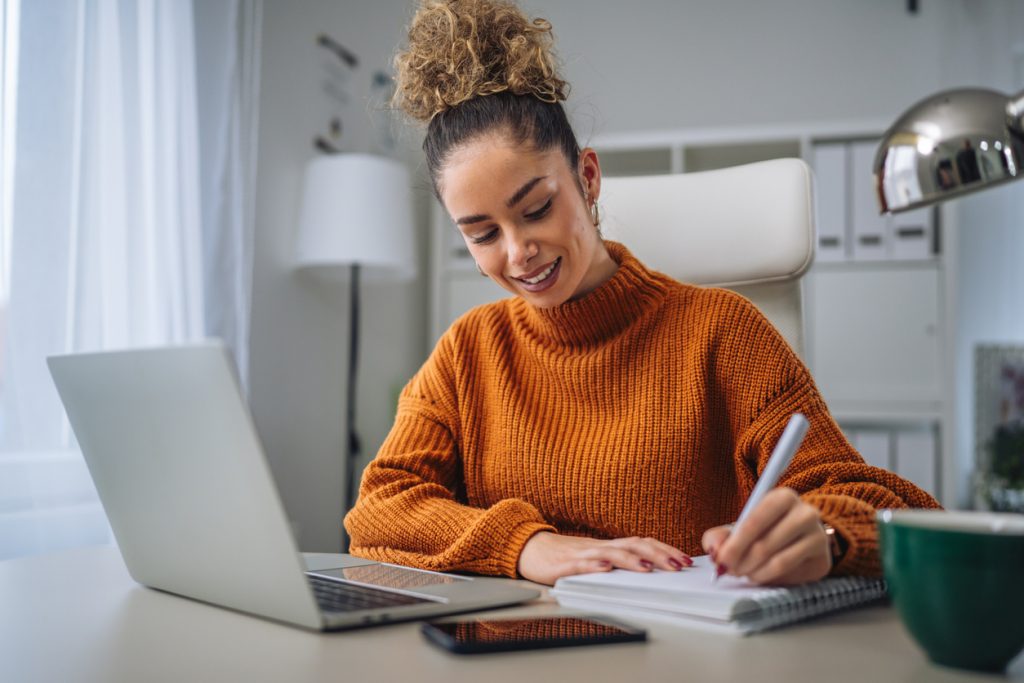 Young professional woman with curly hair bun wearing an orange sweater sitting at her desk, typing on a laptop, and taking notes in a spiral notebook, working remotely from a modern home office