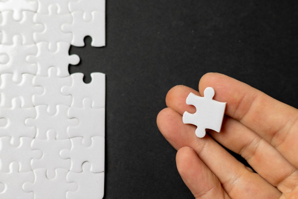 Close-up of a hand holding the missing piece of a white jigsaw puzzle against a dark background. Concept image representing problem solving, finding solutions, business strategy, teamwork, completion and goal achievement.
