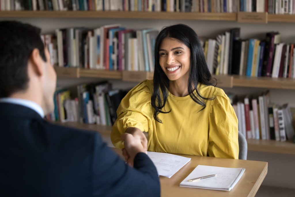 Cheerful Indian student woman talking to interviewer in college library, giving handshake to interviewer, thanking, sitting at table with bookshelves behind