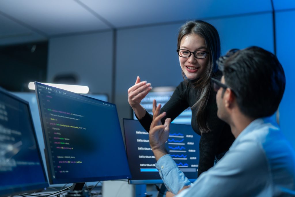 Female programmer gestures while talking through an idea with a smiling colleague at their coding desk, engaged in lively teamwork.